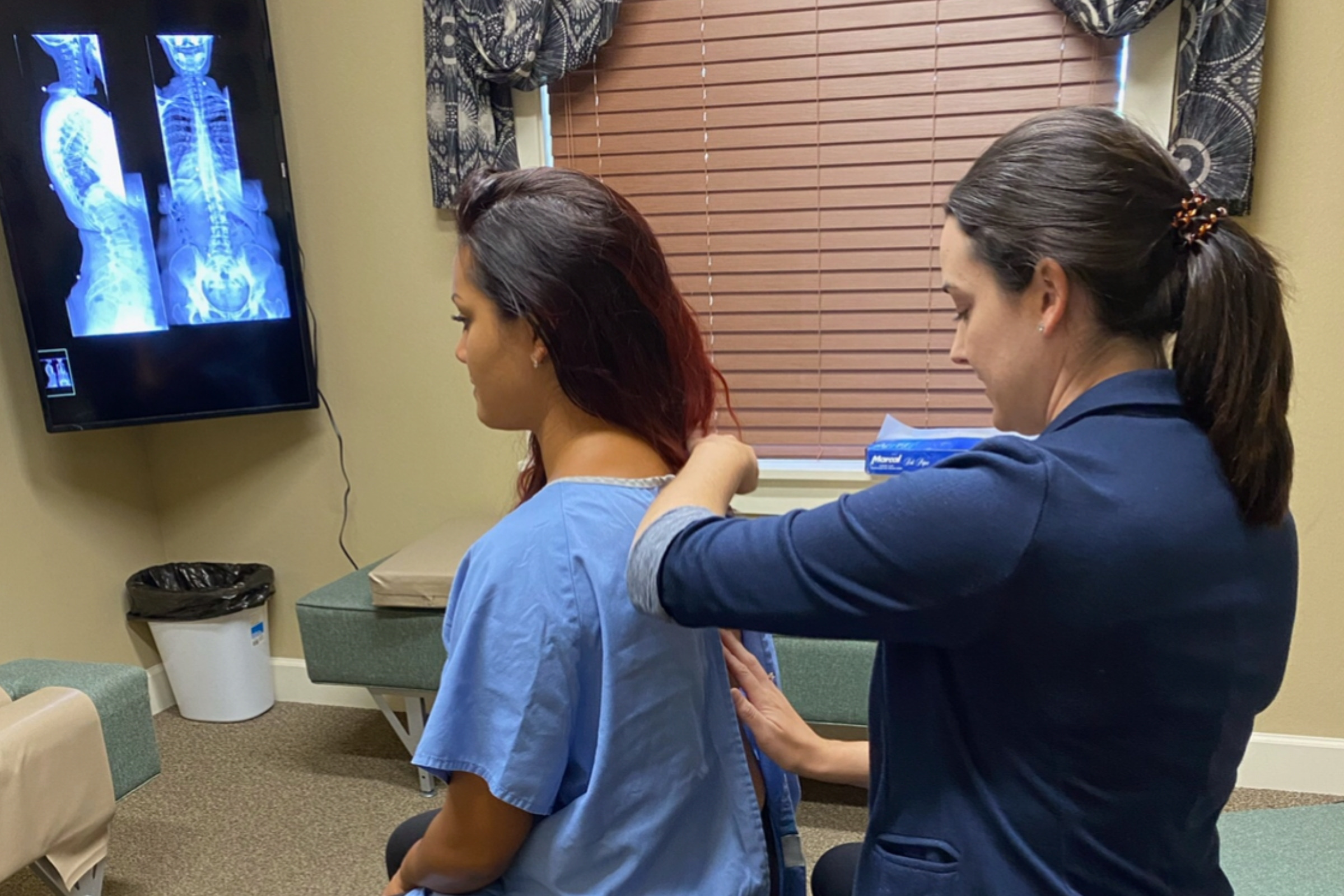Dr. Raquel Heisse examining a seated patient’s spine by hand, with full-spine X-rays lit on the wall behind her